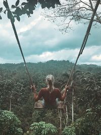 Rear view of woman swinging over trees against cloudy sky in forest