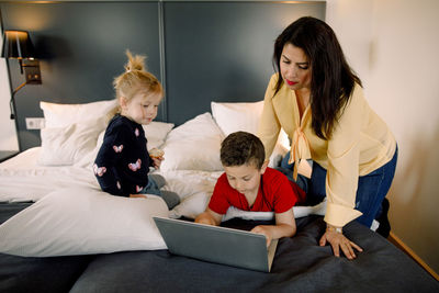 Mother and girl with boy using laptop on bed in hotel room