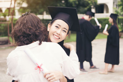 Cheerful woman in graduation gown embracing mother while standing outdoors