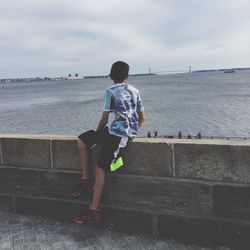 Side view of boy sitting on retaining wall against sky