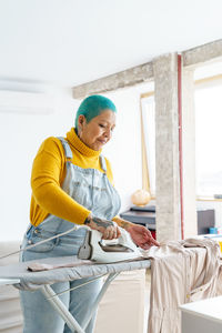 Content female with short dyed hair in overall ironing clothes on board during housework routine in light room at home