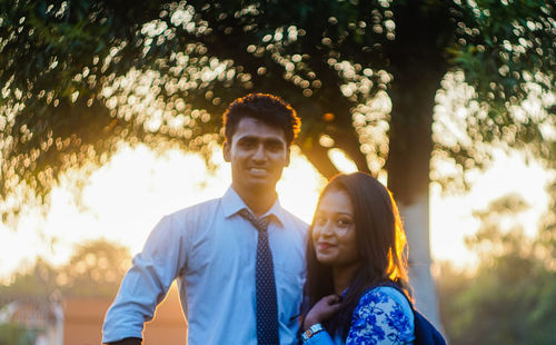 Portrait of happy young couple standing at park during sunset