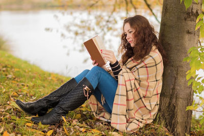Side view of woman reading book while leaning on tree trunk at lakeshore