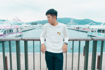 Young man looking away while standing by railing against sea