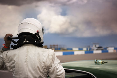 Rear view of man wearing helmet standing by car against cloudy sky