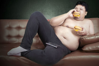 Shirtless man eating burger while leaning on sofa at home