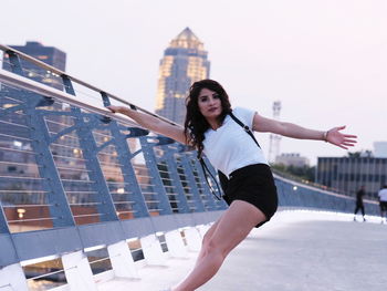 Portrait of mid adult woman holding railing while balancing on footbridge against sky in city during sunset