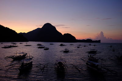 Silhouette outrigger canoes sailing by sea by mountains against sky during sunset