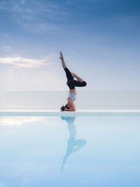 Woman practicing yoga on infinity pool by sea against cloudy sky