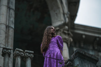 Low angle view of young woman standing against building and looking away 