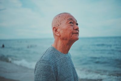 Thoughtful senior man standing at beach against sky during sunset