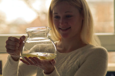 Close-up of smiling woman holding glass jar against window at home
