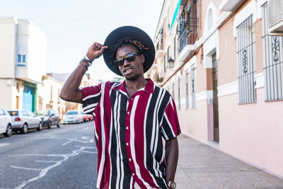 Low angle of confident young african american male in stylish stripped shirt and sunglasses with hat looking at camera while standing against blurred urban street