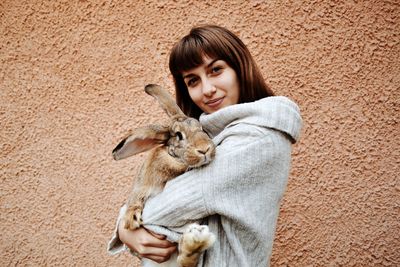 Portrait of woman holding rabbit while standing by wall