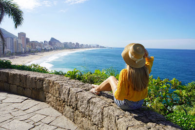 Woman with straw hat sitting on wall looking view of rio de janeiro with leblon and ipanema beaches