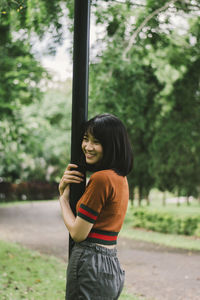 Smiling young woman standing by pole against trees in park
