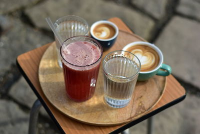 Cappuccino, macchiato, raspberry lemonade and two water glasses on a wooden tray, in a public park