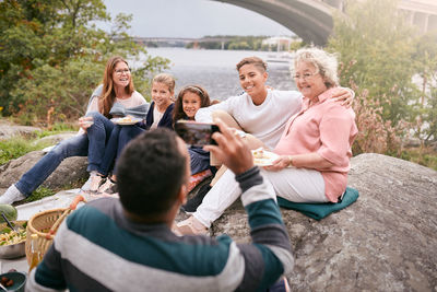 Man photographing happy family with meal on rock in park during picnic
