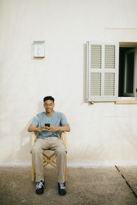 Full length of young man using smart phone sitting on chair against white house