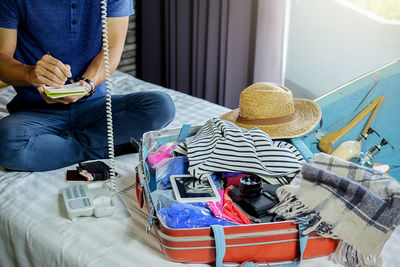 Midsection of man using telephone while packing suitcase on bed at home