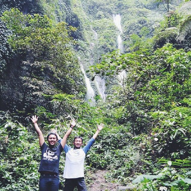 lifestyles, leisure activity, tree, waterfall, water, forest, nature, casual clothing, rock - object, standing, green color, beauty in nature, growth, plant, young adult, vacations, three quarter length, men