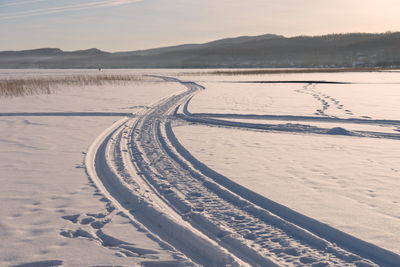 A trail from a snowmobile leads into the distance along the white snow of the frozen lake at sunset.