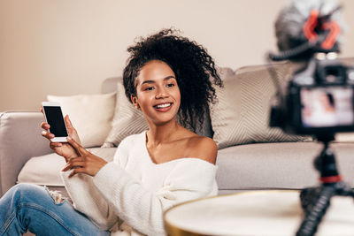 Smiling young woman using phone while sitting by sofa at home
