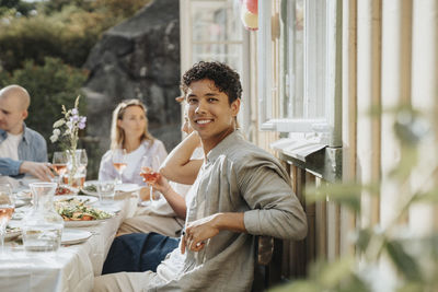 Side view portrait of smiling young man sitting on chair with friends during dinner party at cafe