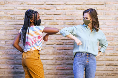 Smiling women wearing mask doing elbow bump standing against wall