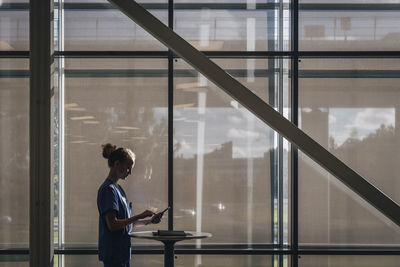 Side view of female nurse using tablet pc while standing by window at hospital