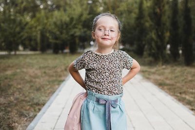 Funny cute child girl 4-5 year old wear glasses and casual clothes hold backpack in park over nature