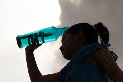 Silhouette of a girl splashing water on head from a blue plastic bottle under sky to cool down