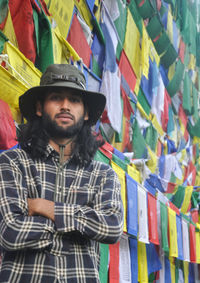 A young guy looking at camera posing with his arms crossed, standing against buddhist prayer flags