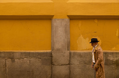 Adult man in hat and coat walking in front of yellow wall on street during sand storm day. madrid.