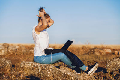 Young woman with laptop sitting on land against sky