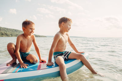 Shirtless boys sitting on paddleboard in sea against sky