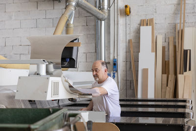 Senior man operating wood cutting machine while sitting at factory
