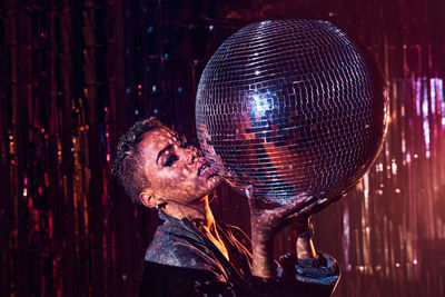 Trendy young ethnic female touching lips while looking at camera and leaning on mirror ball in nightclub
