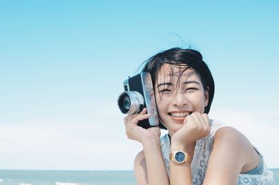 Close-up of smiling young woman holding camera against sky