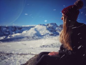 Side view of young woman sitting on snow covered field against blue sky