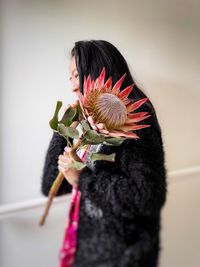 Close-up of woman holding pink king protea flower against white wall.