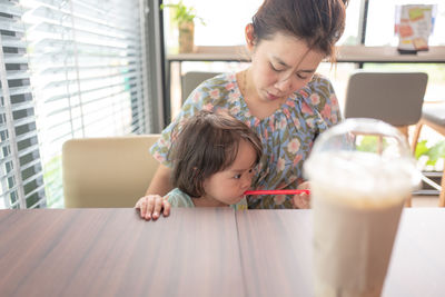 Girl with drinking straw in mouth sitting by mother at table in restaurant 