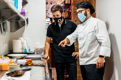 Male cook in medical mask talking to waiter and pointing at prepared delicious dish ready for serving in cafe