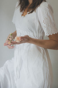 Close up of midsection, a woman holding a pink carnation in her hands while wearing a white dress. 