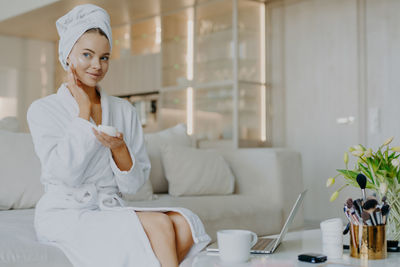 Young woman applying moisturizer while sitting on sofa at home