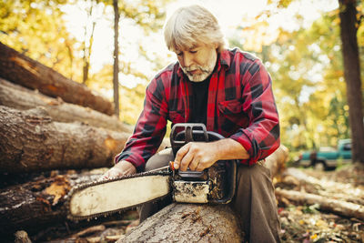 Male lumberjack examining chainsaw while sitting on log in forest