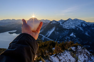 Cropped hand holding snow against mountain during sunset