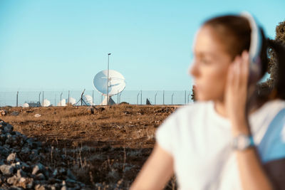 Young woman listening over headphones while sitting on land against sky