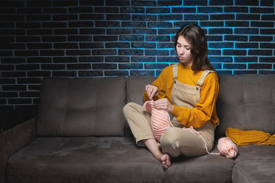 Portrait of cute young caucasian woman sitting on sofa at home and crocheting wool product