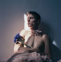Shirtless young man with flowers sitting against wall